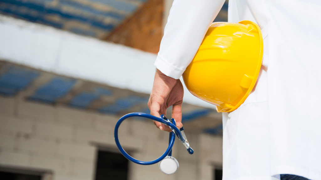 Operatore sanitario con camice bianco tiene in mano un casco da cantiere giallo e uno stetoscopio, simbolo della medicina del lavoro nei luoghi ad alto rischio.